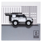 Metal enamel pin of a white Land Rover Defender with white wheels, spare wheel and roof rack. In collaboration with the Lamley Group. On backing card resembling a white shipping container.