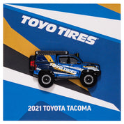 Metal enamel pin of a 2021 Toyota Tacoma with custom livery and Toyo Tires logo. Colors: light and dark blue, yellow, and white. Equipped with black and silver off-road wheels and tires, spare wheel, gas tank, truck bed stroage, roof rack, and off-road lights. On backing card resembling the custom livery with Toyo Tires logo.